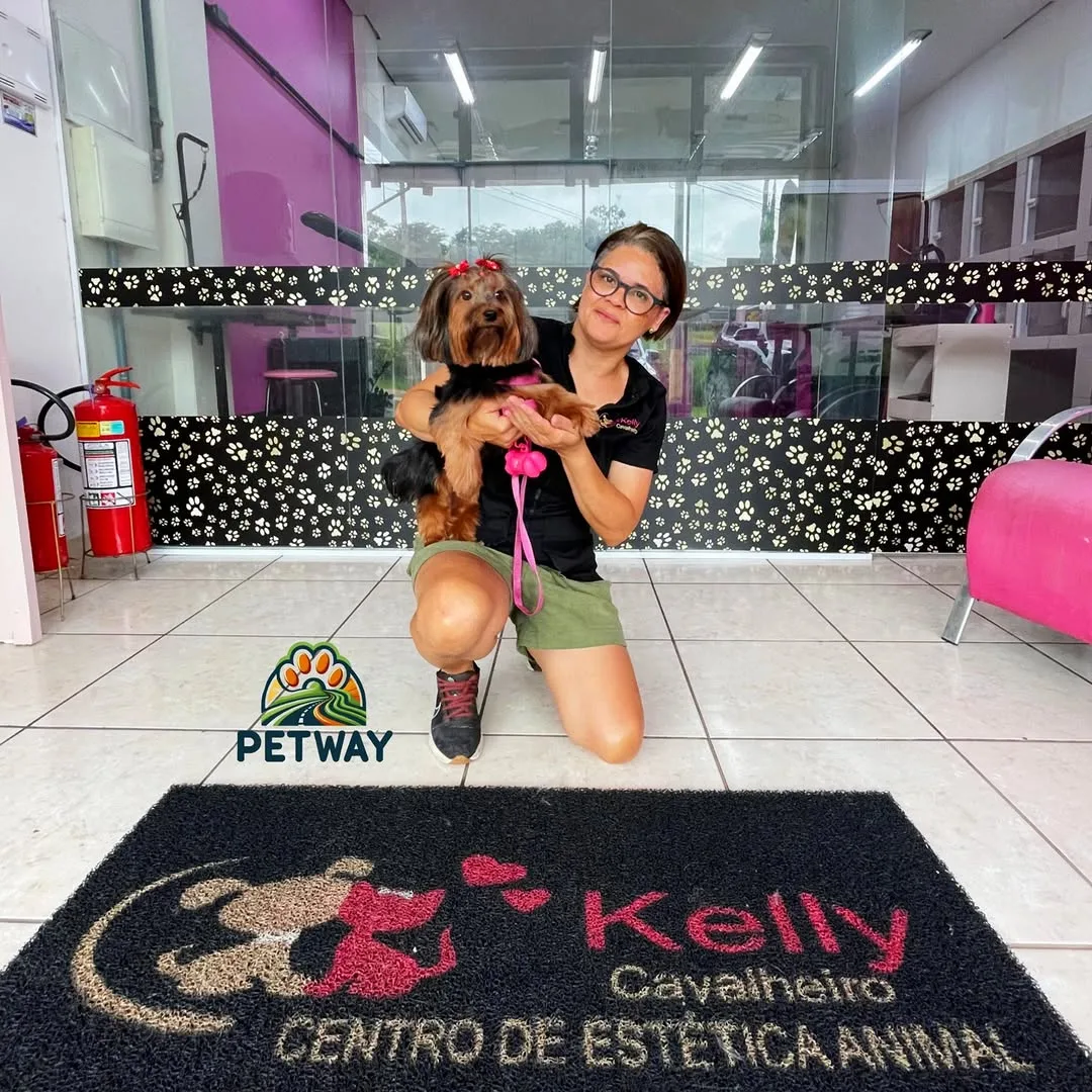 Kelly na recepção do salão, segurando um yorkshire, com a área de banho e tosa envidraçada ao fundo